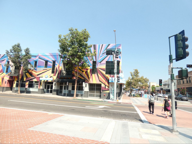 A geometric design mural called 4th Street Market Mural covering an entire building with pinks ranging to purple on the top design and yellow to burnt sienna on bottom with street on side and front.