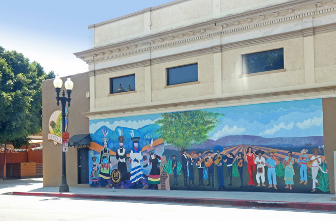 A mural depicting a Chinelos carnival with maskers dressed in traditional costumes and people gathered with musical instruments in front of a field.