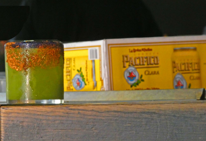 A glass of green michelada with red salt rim sitting next to Cerveza Pacifico Clara carton.
