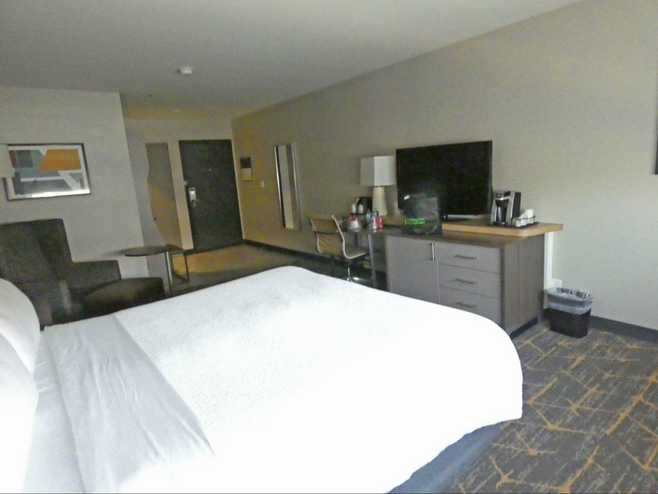 A hotel room with bed, lounge chair, table, desk and dresser with TV above it.