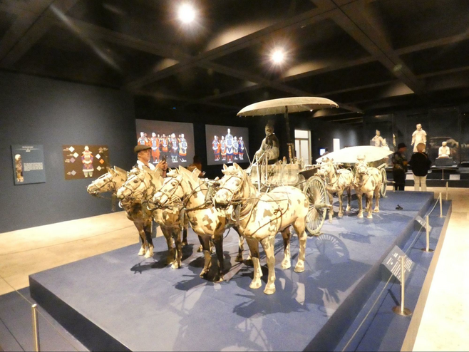 In front, an open chariot drawn by four bronze horses, with a single standing driver and a bronze umbrella on a stand placed next to him. Behnd is "bronze chariot number two," a closed carriage with two seats and an umbrella-like roof, which is also drawn by four bronze horses at Bowers Museum.