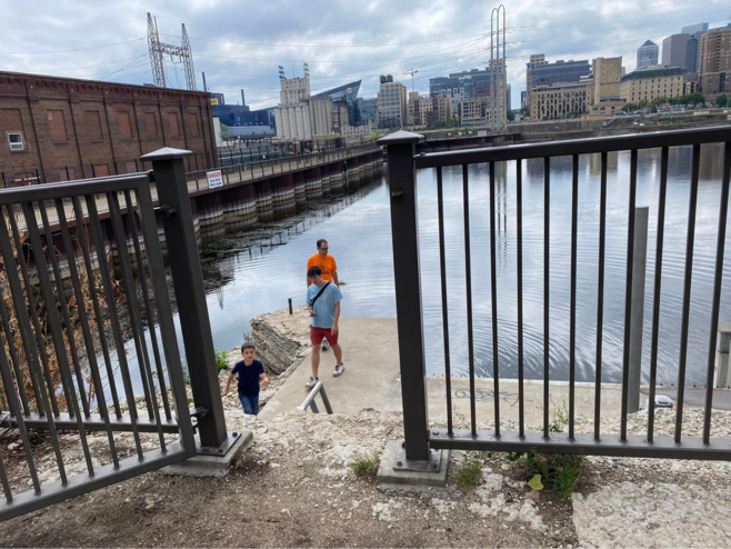 The Mississippi River with the St. Paul skyline behind it. A father and two sons are near the river. There is a gate with a open door leading to the river. 