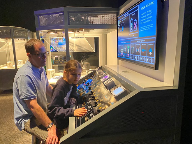 A father and daughter sit at a console at the Science Museum of Minnesota. They are reading about the exhibit. A screen above them explains their mission. 