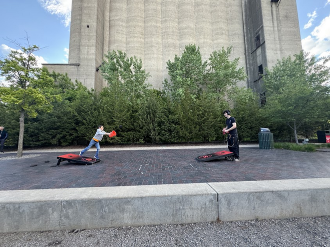 Two boys playing corn hole. One boy is wearing black. The other, younger boy is in a grey sweatshirt holding  a red bucket.   