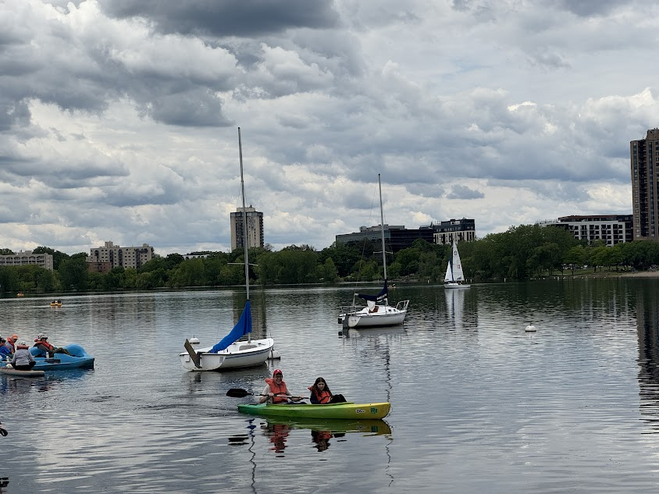Kids kayaking on a lake with green treed shoreline and moored sailboats in the background.