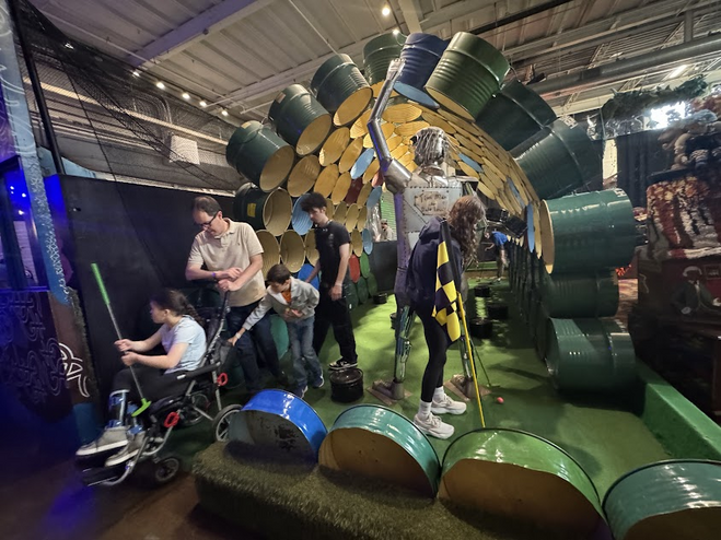 Family playing mini-golf. Father is with a girl in a wheelchair, a girl is putting, and two boys are standing by waiting their turn. 