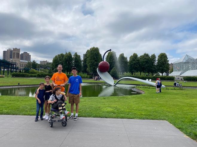 Sculpture of a giant spoon with a cherry on top by a small lake. A family of five is in front of the sculpture, a father with two boys and two girls. One of the girls is in a wheelchair. 