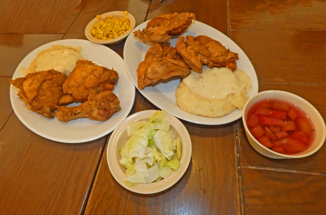 Two plates of fried chicken with mashed potatoes and several bowls of vegetables on table.