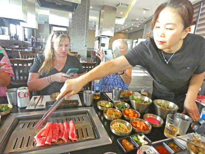 Two women sitting at table with an array of vegetable dishes and BBQ built in while server cooks meat.