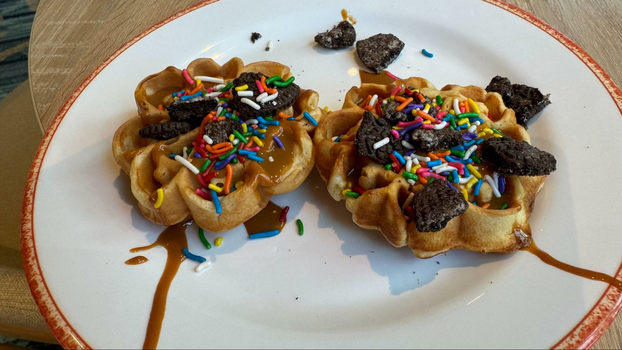 Two waffles with oreos and rainbow sprinkles on a white plate with an orange rim.