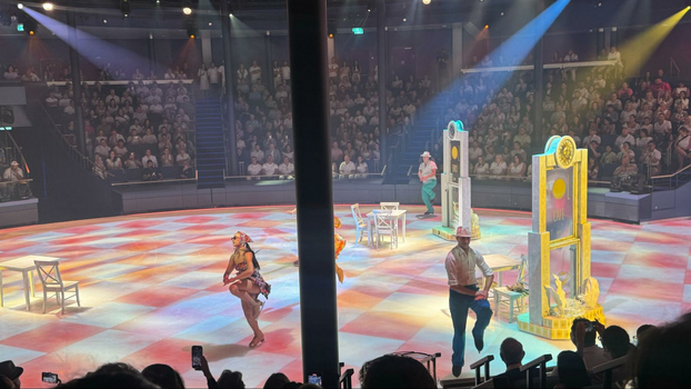 a circular theater with an ice rink in the center with four skaters pressing their hands to their knees next to props of chairs and doors.