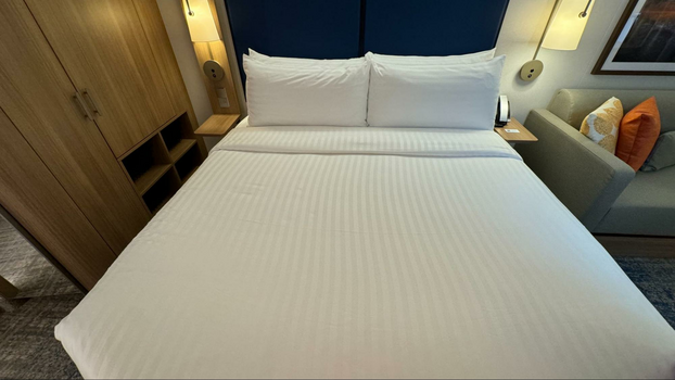Bed with white comforter and four pillows stacked up against a navy blue headboard next to a wooden closet and a beige sofa. 