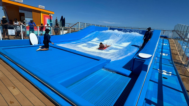 blue water splash pad with a man on a red boogie board next to two employees.