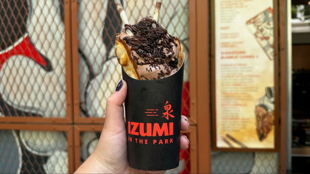 Black ice cream holder with red lettering that says Izumi in the Park with chocolate ice cream with chocolate sprinkles in a bubble cone in front of a store sign