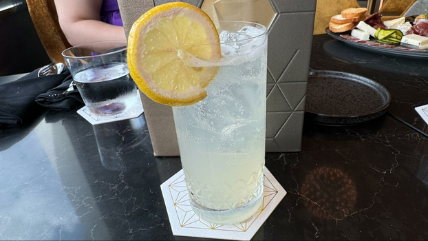 A tall glass of a carbonated beverage with a lemon wedge on top of a white coaster.