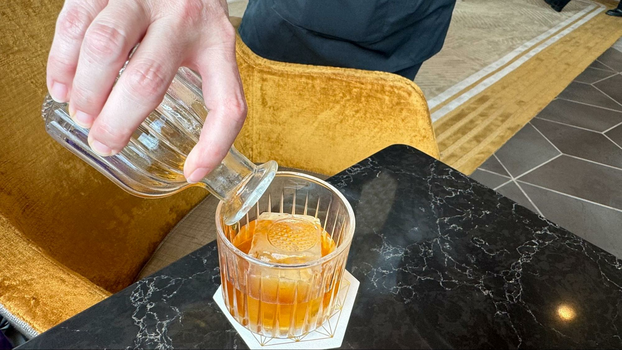 A servers hand pouring brown butter old fashioned into a glass with a stamped ice cube of the geodesic Epcot structure. 