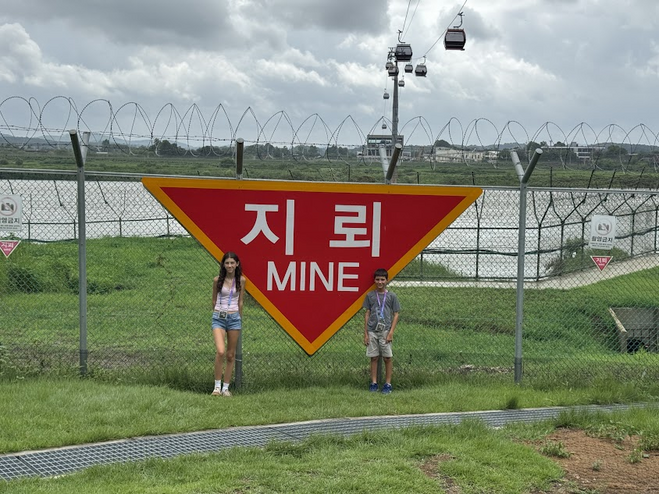 Two children standing in front of a large red sign that says "mine" in white letters in English and Korean. Two children are standing in front of the sign. A barbed wire fence and a river are in the background.  