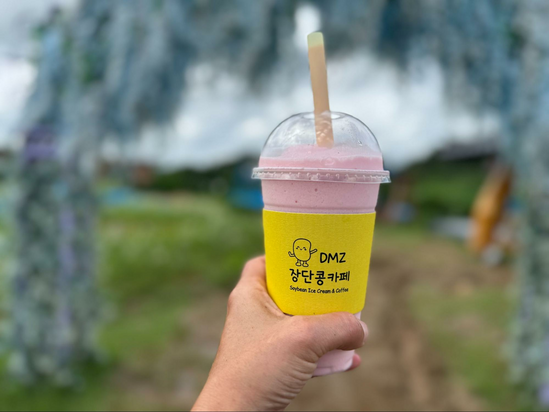 Hand holding a pink smoothie with a yellow sleeve that says DMZ. The smoothie is against a background that includes a park and purple flowers. 
