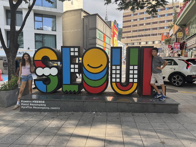 A boy and a girl in shorts standing in front of a colorful sign that says Seoul. 