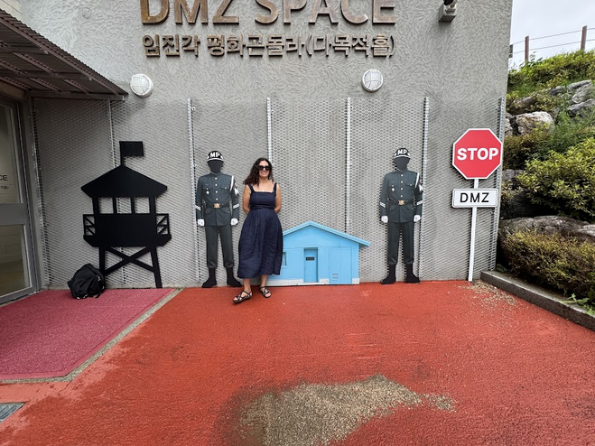 Woman in a black dress and sandals standing in front of a replica checkpoint. There is a red stop sign with a black and white sign reading DMZ, a small light blue hut and two cut-outs of soldiers. 