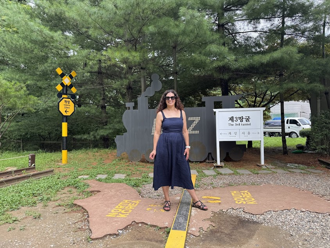 Woman in a black dress and sandals standing over a yellow line with North Korea written on one side and South Korea on the other. There are trees in the background. 