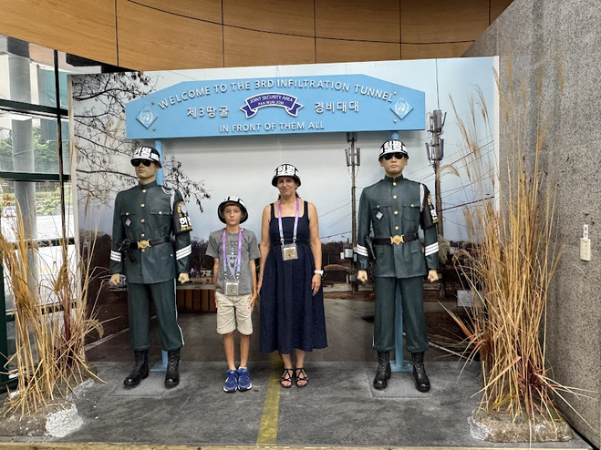 A mother and son standing with two statutes of Korean soldiers. They are wearing helmets. A light blue sign above them reads "Welcome To The 3RD INFILTRATION TUNNEL" in white letters. 