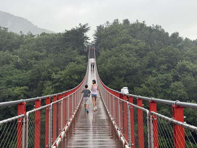 A boy and a girl in shorts walking on a suspension bridge in the mountains. The sides of the suspension bridge are red. 