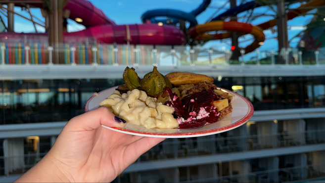 Hand holding a plate with macaroni and cheese, beets & feta, vegetables and yuca against a window overlooking the water park on Royal Caribbean's Star of the Seas