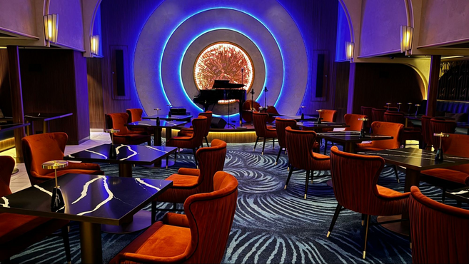 A room featuring orange velvet chairs and black chic tables against a patterned carpet floor in front of a stage with three circles of blue lights and a piano