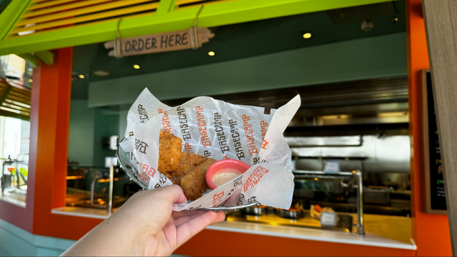 Hand holding a basket of chicken tenders with a side of honey mustard in a red cup in front of Basecamp eatery aboard Star of the Seas