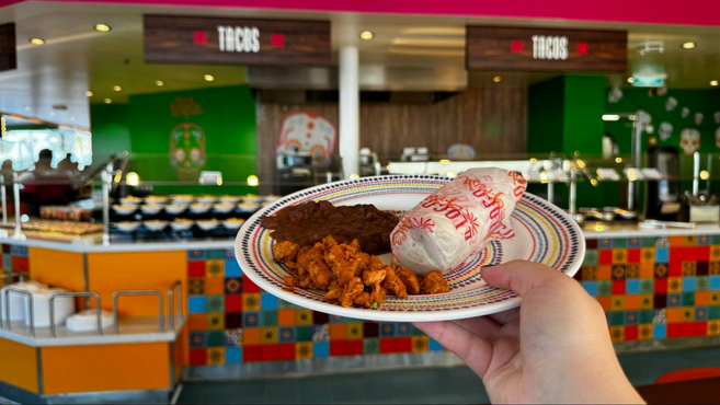 Hand holding a colorful plate containing a burrito, chicken and black beans, in front of a rainbow tile decoration at the El Loco Fresh restaurant on Star of the Seas