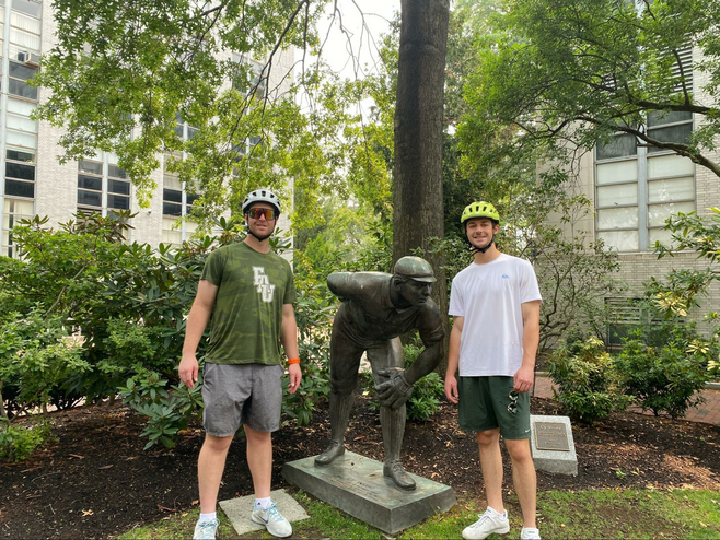 Two young guys in bike helmuts standing next to a concrete statue of Cy Young.