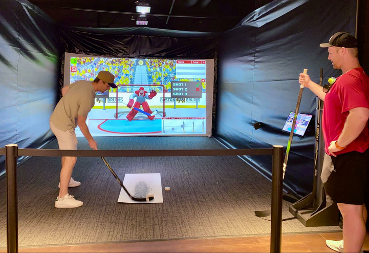 A young guy holding a hockey stick hitting the puck into a simulator screen as another young guy looks on.
