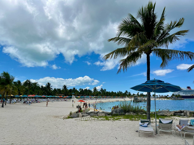The beach (dotted with colorful beach umbrellas and palm trees) at Disney Cruise Line's private island destination in the Bahamas, Castaway Cay. The cruise ship Disney Wish is pictured in the distance.