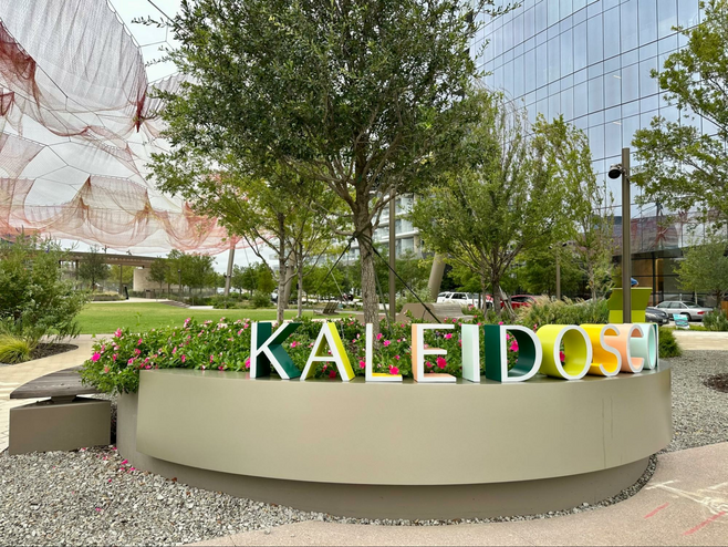 Rainbow-hued Kaleidoscope Park sign in Frisco, Texas, with a large-scale fiber arts installation visible in the background.