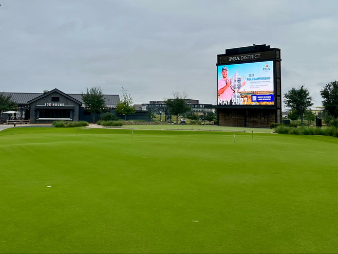 Giant TV screen at the PGA District in Frisco, Texas, with a putting green in the foreground where adults on a Frisco sports getaway can play.