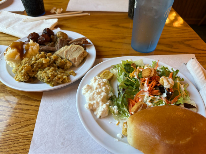 Two plates filled with buffet food - the front has salad, cottage cheese and a roll and the plate behind has turkey, roast beef, mashed potatoes and stuffing. A glass of water also sits on the table.
