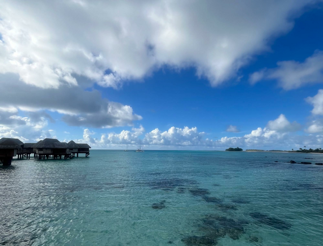 Bungalows appear over the crystal-clear ocean waters of French Polynesia. The sea is so clear you can see the coral below, clouds hover in the distance against a bright blue sky.