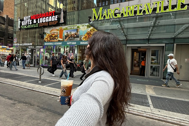 Entrance to Margaritaville Resort, Times Square, NYC. A brunette woman with long hair is taking a selfie while looking away and holding a coffee drink.