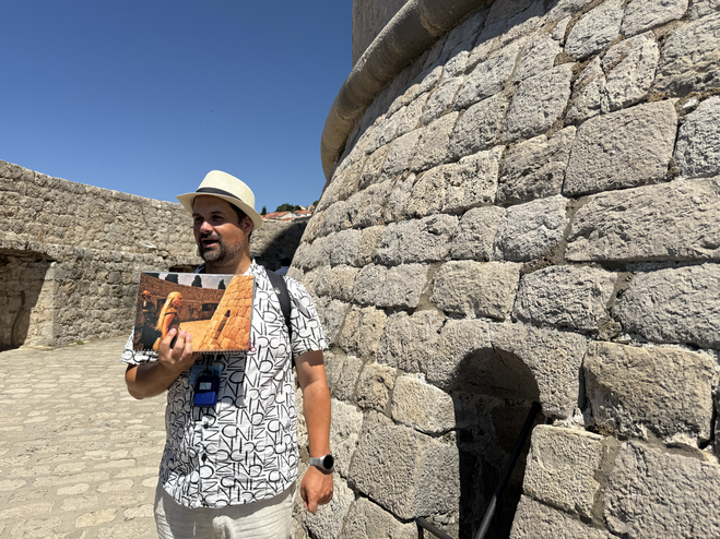 Game of Thrones Tour guide standing next to old Dubrovnik city wall with scene card from Game of Thrones TV show.