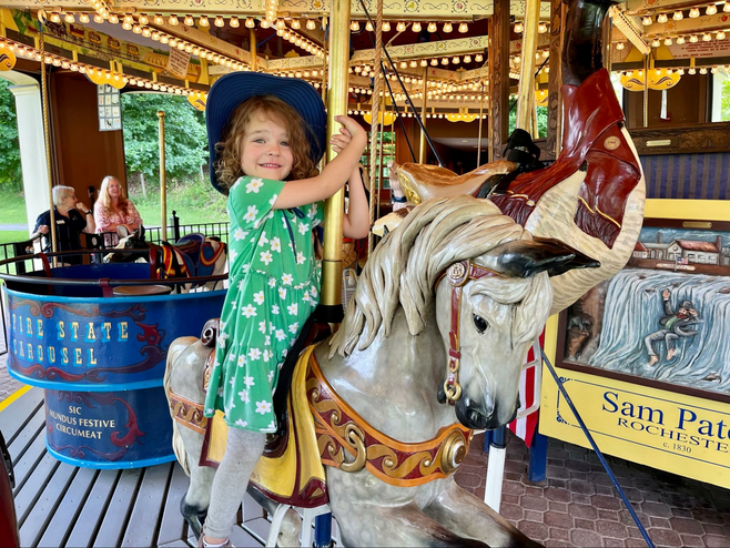 A young girl perches on a hand-carved, hand-painted regal grey carousel horse on the Empire State Carousel at Fenimore Farm & Country Village in Cooperstown, New York. 