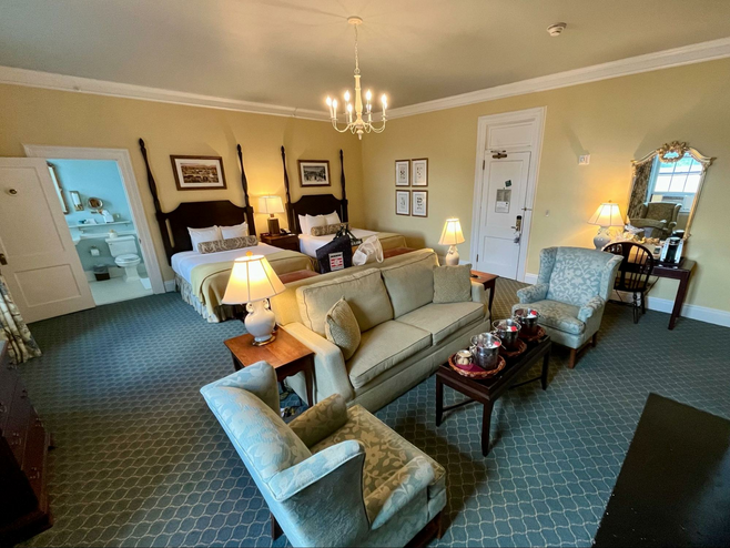 A spacious suite at The Otesaga Resort Hotel, a historic luxury hotel in Upstate New York. Pictured is a huge hotel room with artwork on stylish yellow walls, two queen-sized beds, a couch and comfortable chairs, a table, desk, upscale blue patterned carpeting and a bathroom. 