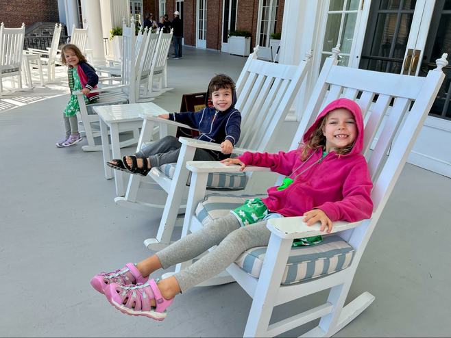 Three children sit in white wooden rocking chairs on the expansive veranda of The Otesaga Resort Hotel in Cooperstown, New York. 