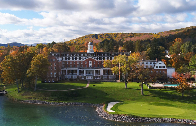 An aerial photo of The Otesaga Resort Hotel on Cooperstown, New York, as seen from the lake in fall. The hotel is red brick and regal, with an expansive veranda. A swimming pool, golf course and lawn can be seen, and fall trees in shades of orange can be seen in the background. 