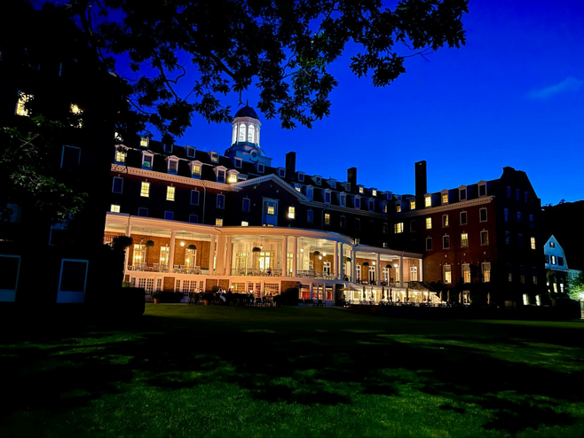 The historic grand red brick exterior of The Otesaga Resort Hotel in Cooperstown, New York, as seen lit-up from the lakefront at night. The resort's iconic veranda is particularly well-lit and visible.