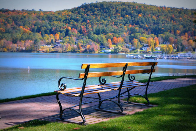 A sunlit bench is pictured on a crisp fall morning facing the lake outside The Otesaga Resort Hotel in Cooperstown, New York. Trees dotted with orange and yellow can be seen across the lake. 