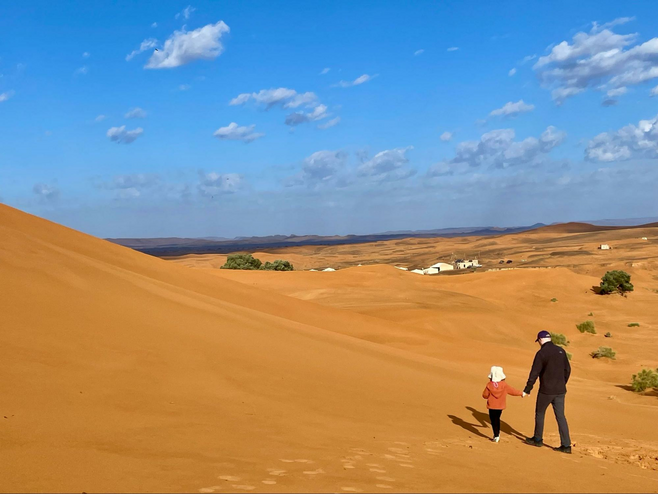 A father and daughter walk amidst the towering ochre-colored desert dunes of Morocco's Sahara.