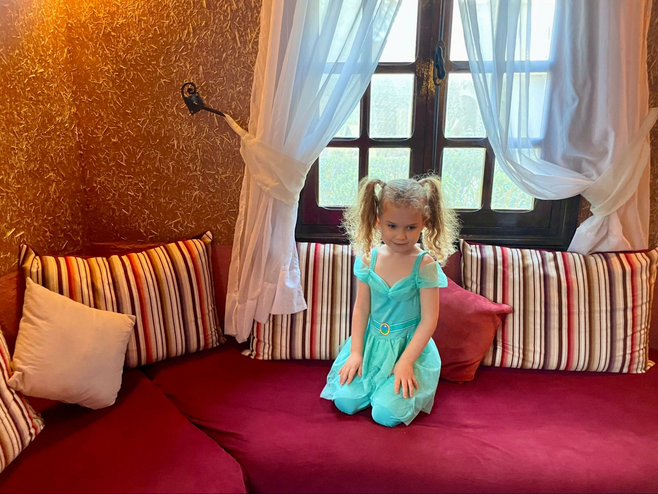 A young girl dressed as Princess Jasmine from the Disney film "Aladdin" sits on a plush couch near a window.