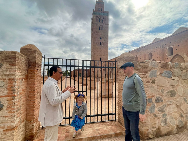 A local guide stands before a famous mosque in Marrakech, explaining its history to a male tourist as a young girl plays.