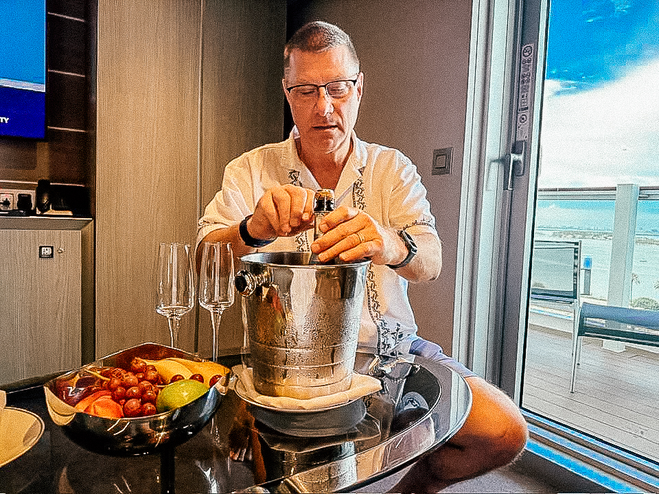 Man with white shirt and glasses in front of coffee table opening champagne